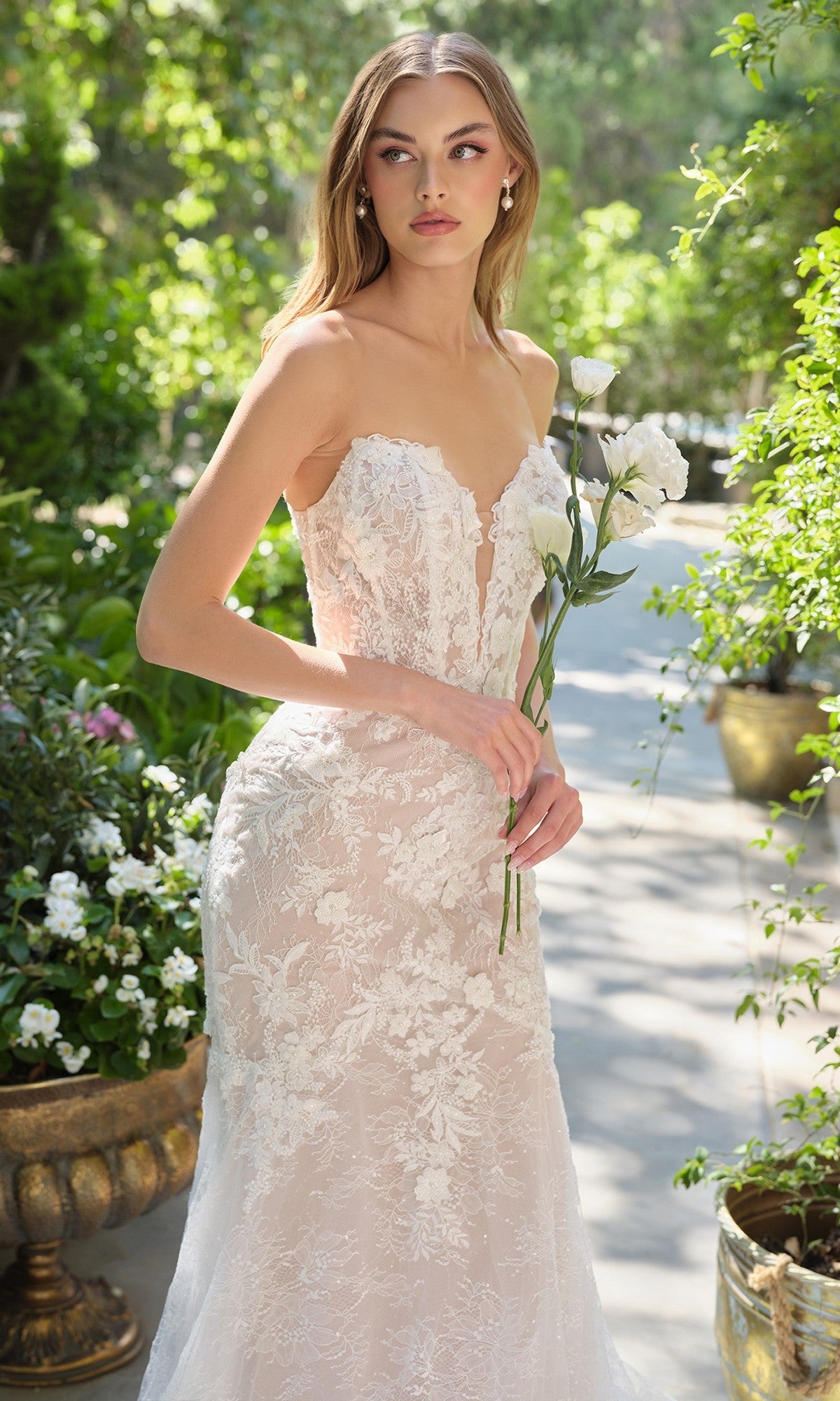 A woman stands outdoors surrounded by greenery and potted plants, holding white flowers and wearing the Andrea and Leo WL072 long wedding dress, a strapless lace mermaid bridal gown.